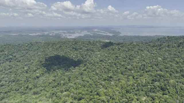 Drone lowers from high over Brownsberg Hill looking out at lake with forested islands on partially cloudy day in Suriname