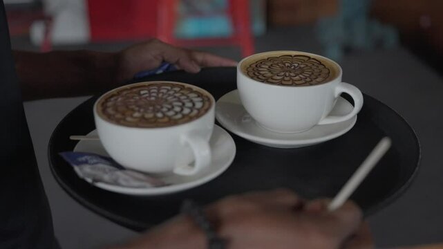 Closeup of a male waiter carrying tray of two floral designed latte mugs in a cafe