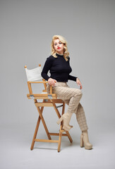 Beautiful girl with blond hair in a black sweater and brown pants posing in a studio on a gray background.