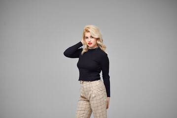 Beautiful girl with blond hair in a black sweater and brown pants posing in a studio on a gray background.