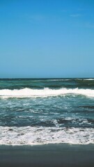 Waves crashing against the shore with a clear blue sky in the background at Parangtritis Beach, Yogyakarta.