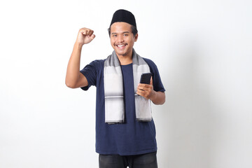 Fototapeta premium Portrait of excited Asian muslim man wearing sarung scarf and skullcap, raising his fist, celebrating winning game or getting good news on his mobile phone. Isolated on white background