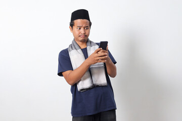 Portrait of upset Asian muslim man wearing sarung scarf and skullcap, holding mobile phone and showing shocked expression. Isolated on white background