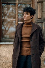 Fashionable young man in brown coat and black turtleneck sweater standing in front of window with old wooden door