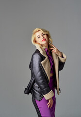 Fototapeta premium A beautiful girl with blond hair in a purple dress stands on a gray background. She is wearing a black jacket with a fur collar.