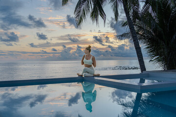 Young woman in a white clothes doing yoga on the beach near  swimming pool early morning in the summer. Sunrise sky in the background .