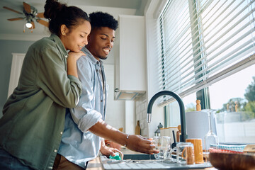 Young black man washing dishes while being at home with his girlfriend.