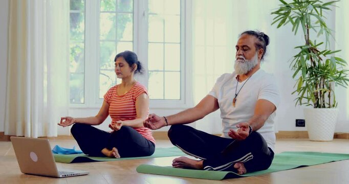 Indian Asian mid aged couple taking an online yoga or meditation class together on a laptop, seated on a yoga mat in their modern home, eyes closed, yog mudra with hands