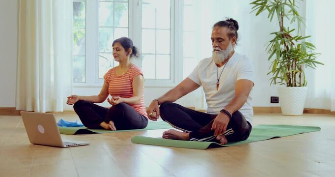 Indian Asian mid aged couple taking an online yoga or meditation class together on a laptop, seated on a yoga mat in their modern home, eyes closed, yog mudra with hands