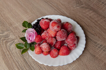 Frozen strawberries with a pink rose on a white plate, cozy food on a wooden table