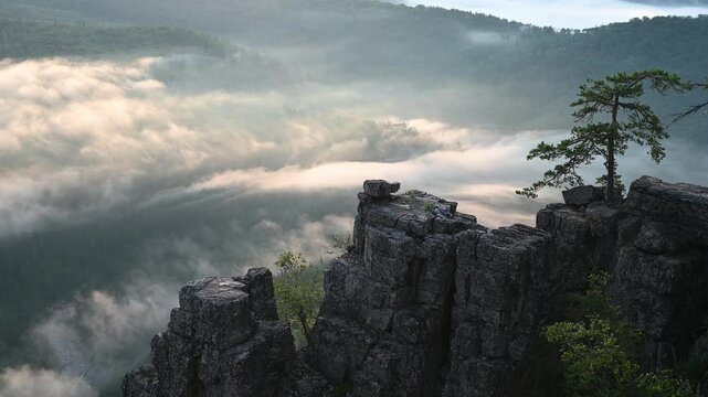 Clouds flow over a mountain valley, creating a stunning view from a high cliff surrounded by lush greenery and rock formations. Ural mountains landscape