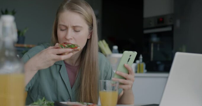 Cheerful young woman using smartphone messaging in social media and having lunch in kitchen at home. Modern technology and youth lifestyle concept.