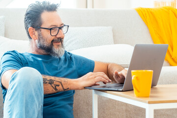 Adult man working remotely from home on his laptop, comfortably seated on the couch, browsing the...