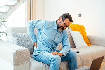 Fototapeta premium Man sitting on a sofa at home, holding his lower back with a pained expression from bad posture during a work break, reflecting stress, poor health, and aging-related physical discomfort