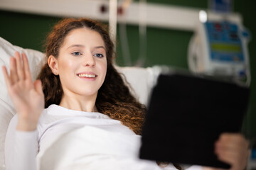 Young woman, sitting in her hospital room with a tablet in hand, waves cheerfully to the camera, maintaining a virtual connection with friends and family, bringing smiles and positivity to her