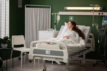 Sick female patient in hospital ward lies in hospital bed looks at window while recovers at hospital.