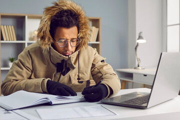Freezing African American man in outerwear working at laptop in office. Businessman wearing warm...
