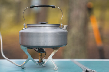 A stainless steel kettle sits on a camping stove, ready for use in a peaceful outdoor environment filled with trees