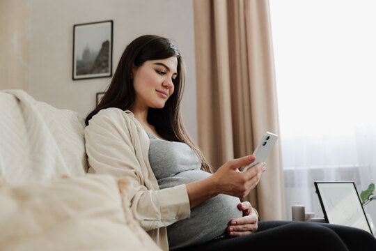 Beautiful smiling pregnant woman with baby belly use mobile cell phone, do online shopping, browse internet, watch podcast while sitting on sofa at home. Having doctor video call medical appointment - Powered by Adobe