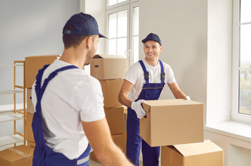 Moving service. Cheerful employees of transportation and delivery company carry cardboard boxes together. Male loaders in overalls are talking cheerfully while packing cardboard boxes in empty office.
