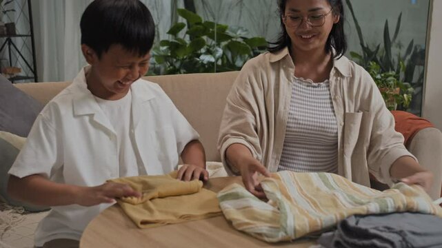 Tilt down shot of cheerful Asian teenage son helping mother to fold laundry after wash in living room