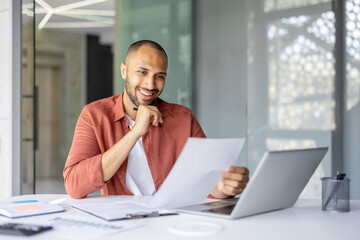 Man wearing casual shirt smiles while reviewing documents at desk. He uses laptop for analysis, portraying successful work environment, professionalism, and confidence in business setting.