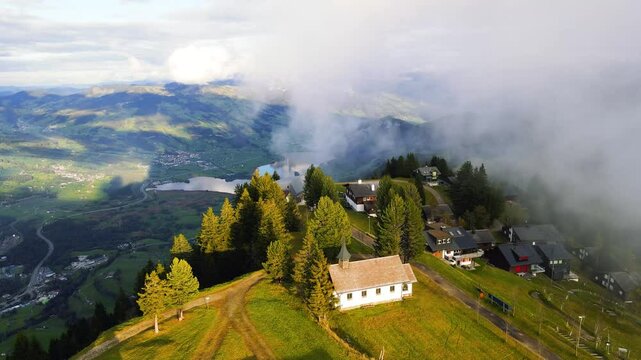 spectacular panoramic shot of a small chapel on the summit in Switzerland overlooking Lake Lauerz surrounded by misty clouds above a lush green valley with clusters of mountain cabins and drifting fog