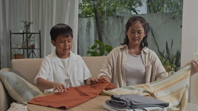 Wide shot of cheerful Asian mother demonstrating teenage son how to fold clothing while sitting on sofa in living room