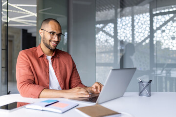 Man smiling while working on laptop in modern office setting. Casual attire with glasses adds to positive work attitude. Shows productivity, business success, technology, and professional lifestyle.