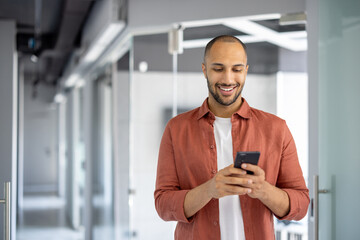 Confident young professional enjoying moment while checking smartphone in modern office hallway. Casual yet focused, he engages with tasks using phone, embodying work-life balance.