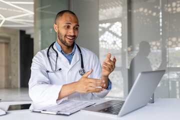 Smiling doctor with stethoscope uses laptop for virtual consultation in modern office. Engages in...