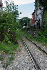 Fototapeta premium A narrow railway track surrounded by dense greenery, running between buildings and trees under a partly cloudy sky.