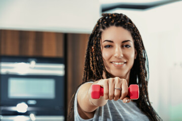 Young woman exercising at home with dumbbells is smiling at came