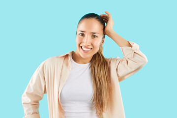 Portrait of a young brunette woman wearing casual clothes standing holding head thinking about something and looking confused at the camera isolated on studio blue background. People emotions concept