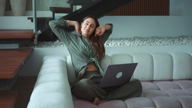 Remote work balance young adult woman relaxes on couch at home taking break from typing on laptop keyboard. Comfortable remote working in living room caucasian female pauses her work on laptop.
