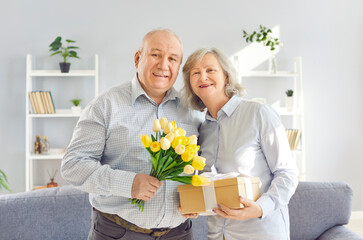 Portrait of an family elderly couple holding gift and a bouquet of flowers, celebrating a holiday at home. They are happy and surrounded by a warm, festive atmosphere, delighted and rejoicing gifts.