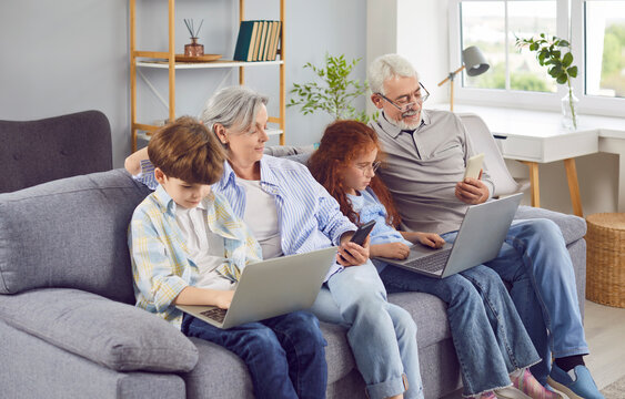 Happy grandparents with grandchildren girl and boy sitting on sofa using modern laptop and smartphones together. Smiling seniors resting on the couch at home with kids. Family and technology concept. - Powered by Adobe