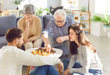 Happy old parents and their adult children are spending time together, sitting in the living room and playing chess on a small table. Family, leisure concept