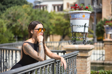 Young woman, brunette, beautiful, very tattooed, with black overalls and glasses with orange lenses, serious and worried, leaning on a railing. Concept tattoos, nonconformism, rebel, trend, fashion.