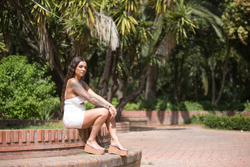 Naklejka premium A young, dark-haired, beautiful woman, heavily tattooed, in a white dress, sitting on a stone bench with a serious and defiant look. Tattoos concept, nonconformism, rebellious, trend, fashion.