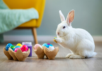A white rabbit curiously examines a colorful Easter egg next to decorated eggs inside a cozy indoor setting