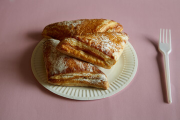 Triangles of puff pastry with sugar sprinkle on a disposable plate with a disposable fork on a pink background, fast food, street food.