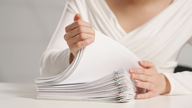Woman sitting at office desk searching through overload stack of paper documents with colorful paper clips. Bureaucracy business concept.