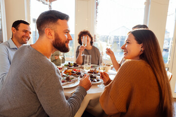 A married couple and their big happy family at a festive table in the dining room. A young man and a woman look at each other in love. People laugh and raise glasses of champagne at home.