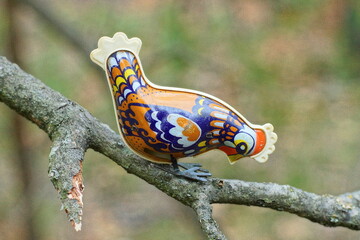 one small beautiful multicolored toy iron painted children's figurine with a white comb and black paws chicken on a tree branch during the day outside