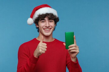 Young caucasian man wearing christmas hat standing over isolated blue background pointing thumb at green screen phone, Generative AI