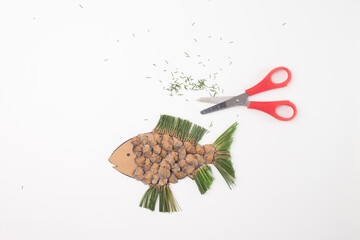 Fish shape made of pine cones and pine needles, white background. red handled scissors next to it, top view, step in DIY tutorial,