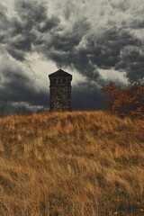 Fototapeta premium Old stone tower on autumn hill with dramatic stormy sky. Gothic architecture landscape. Mysterious rural scene. Moody nature photography with copy space