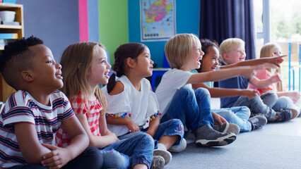 Group Of Smiling Elementary School Pupils Sitting On Floor In A Line Listening To Teacher 