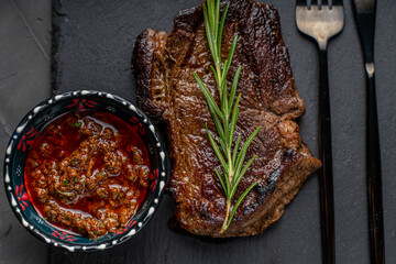 Grilled Marbled Beef Steak with a sprig of rosemary on a mica board with sauce and Argentinian Chimichurri sauce in a bowl on a dark background